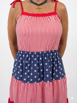 A close-up product view focusing on the fabric details of a 4th of July themed dress. The image showcases the transition from the red and white vertically striped top bodice to the gathered navy blue tier adorned with white stars. A gold star pendant necklace is visible at the neckline against a clean white background.