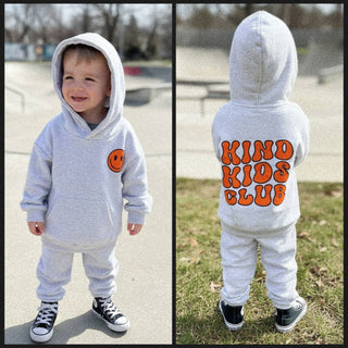 A toddler wearing a light gray hooded sweatshirt and matching sweatpants in an outdoor park setting. The front view shows a small orange smiley face on the chest, and the back view shows the words 'KIND KIDS CLUB' in large orange letters with a darker outline.