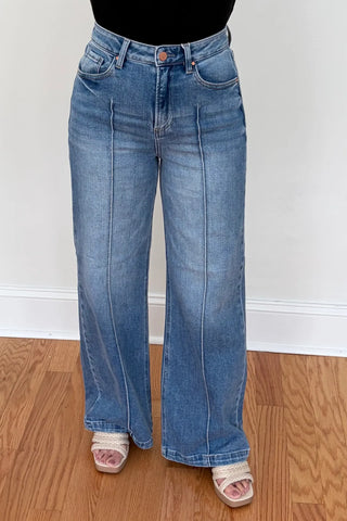 A person wearing light blue wide-leg denim jeans with a black top and beige slide sandals, standing on a wooden floor against a white wall.