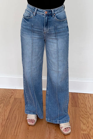 A person wearing light blue wide-leg denim jeans with a black top and beige slide sandals, standing on a wooden floor against a white wall.