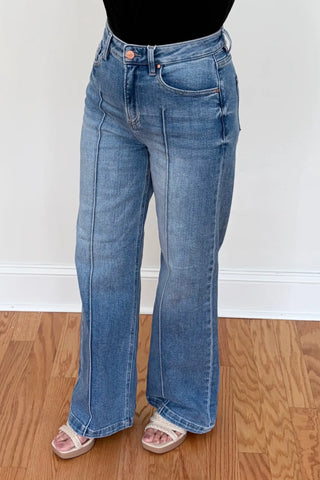 A person wearing light-wash blue wide-leg jeans with a black top and clear slide sandals, standing on a wood floor against a white wall.