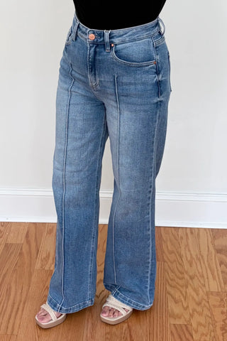 A person wearing light-wash blue wide-leg jeans with a black top and clear slide sandals, standing on a wood floor against a white wall.