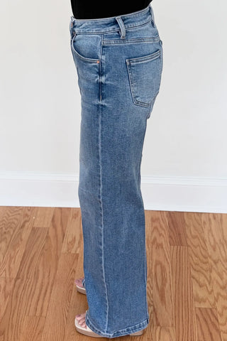 A person wearing a pair of light blue denim flare jeans, shown from the waist down, standing on a wooden floor against a white wall.