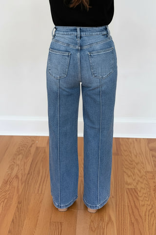 A person shown from the back wearing light-wash blue wide-leg jeans, standing on a wood floor against a white wall.