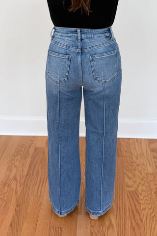 A person shown from the back wearing light-wash blue wide-leg jeans, standing on a wooden floor against a white wall.