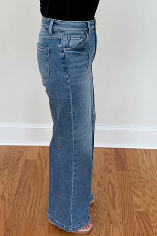 A person wearing a pair of medium-wash blue flare jeans with a black top, standing on a wooden floor against a white wall.