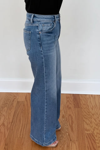 A person wearing a pair of medium-wash blue flare jeans with a black top, standing on a wooden floor against a white wall.