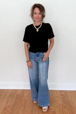 A woman stands in a room with light wood floors and a white wall, wearing a black short-sleeve top, light-wash blue flare jeans, a pearl necklace, and white slide sandals.