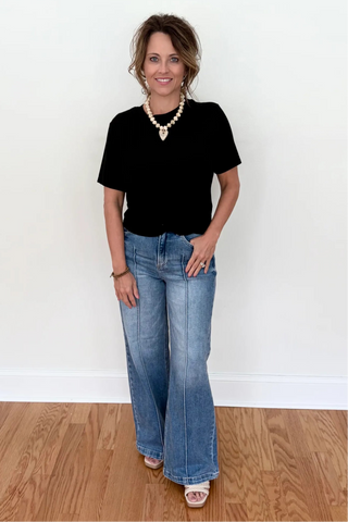 A woman stands in a room with a white wall and wood floor, wearing a black short-sleeve top, light-wash blue flare jeans, a gold beaded necklace, and white slide sandals.