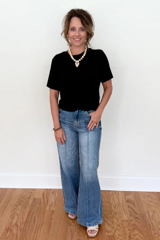 A woman stands in a room with a white wall and wood floor, wearing a black short-sleeve top, light-wash blue flare jeans, a gold beaded necklace, and white slide sandals.