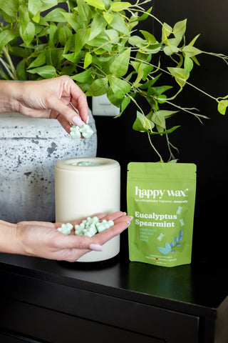 A person's hands are shown holding and sprinkling light green wax melts next to a white jar and a green Happy Wax Eucalyptus Spearmint pouch on a black table.