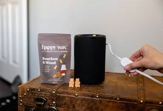 A person's hand holds a white cord near a black wax warmer next to a bag of Happy Wax Bourbon & Wood scented wax melts, with three small wax melts on a brown leather suitcase.