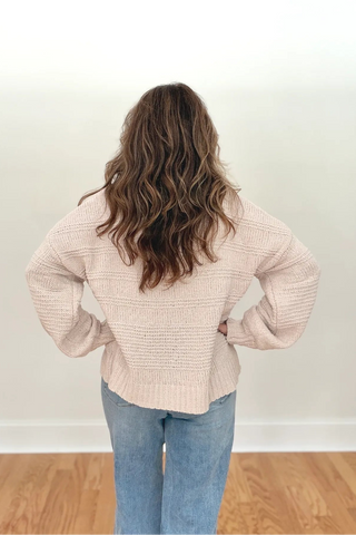 A woman viewed from behind wearing a light beige textured knit sweater and blue jeans, standing on a wooden floor against a plain white wall.