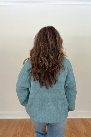 A person viewed from behind wearing a light blue knit sweater and light blue jeans, standing in a room with a white wall and wood floor.