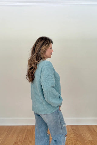 A woman wearing a light blue knit sweater and light-wash blue jeans, standing in profile against a plain off-white wall with a wooden floor.