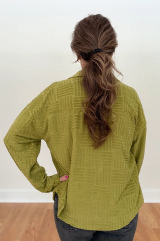 A woman viewed from behind wearing a textured olive green cardigan with a geometric basketweave pattern, paired with dark gray jeans. She is standing in a room with a white wall and light wood floor.