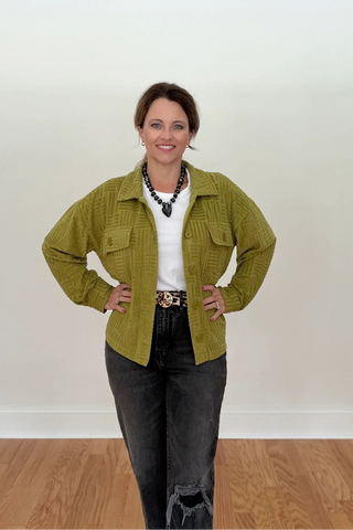 A woman stands in a room with a white wall and wood floor, wearing an olive green textured button-up jacket over a white top with dark gray jeans.