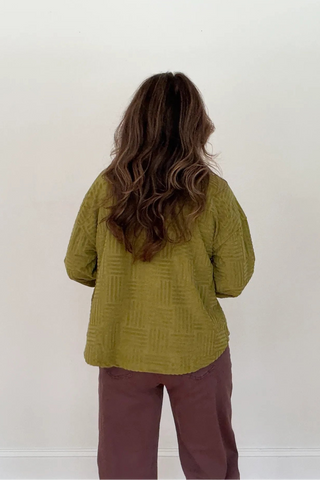 A woman viewed from behind wearing a textured olive green long-sleeve top and mauve-brown pants, standing against a plain off-white wall.
