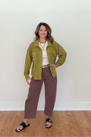A woman stands in a room with a white wall and light wood floor, wearing an olive green utility-style jacket over a cream top, mauve-brown wide-leg pants, and black slide sandals.