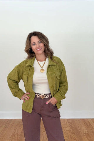 A woman modeling a textured olive green zip-up jacket over a cream top and brown pants, standing against a plain white wall with wood flooring.