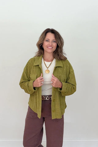 A woman modeling a textured olive green quilted jacket over a cream top and brown pants, standing against a plain white wall.