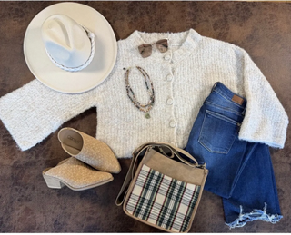 A styled flat-lay outfit arrangement on a brown textured surface, featuring a cream knit cardigan, blue jeans, beige heeled mules, a white wide-brim hat, a plaid shoulder bag, and small accessories.