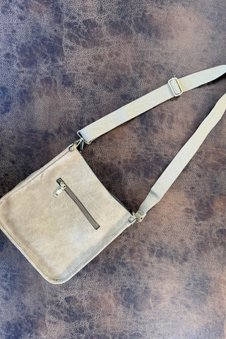 A distressed beige leather crossbody bag with a matching fabric strap and silver-tone hardware, laid flat on a textured brown surface.