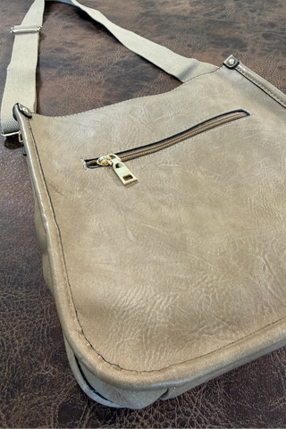 A light beige faux-leather shoulder bag with a front zippered pocket and adjustable strap, shown on a dark brown textured surface.