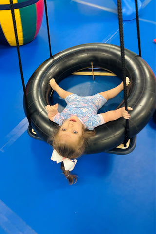 A toddler girl wearing a blue patterned romper lies inside a black tire swing on a bright blue indoor playground floor.