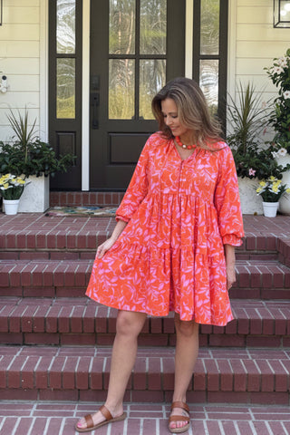 A woman wearing a bright coral-orange and white floral print dress stands on the brick steps of a yellow house porch.