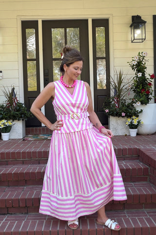 A woman wearing a pink and white vertically striped sleeveless maxi dress with a matching belt stands on brick steps in front of a house with yellow siding and dark doors.