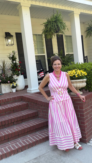 A woman wearing a pink and white vertically striped sleeveless maxi dress with a matching belt stands on the front steps of a house with white columns and brick steps.