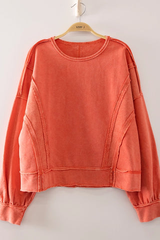 A coral orange long-sleeve cropped sweatshirt with unique diagonal seam details, hanging on a wooden hanger against a plain light gray wall.