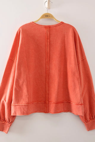 A coral orange long-sleeve top hanging on a wooden hanger against a plain light gray wall. The back of the garment features a central seam running from the neckline to the hem, with a raw, frayed edge.