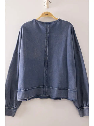 A washed blue denim long-sleeve top hanging on a wooden hanger against a plain light gray wall. The back of the garment features a central seam and raw, frayed hems.