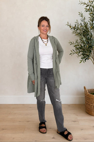A woman stands in a bright room wearing a sage green open-front jacket over a white top, gray jeans, and black slide sandals. A potted olive tree in a woven basket sits to the right against a light beige wall.