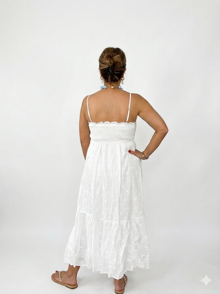 Rear view of a women's white tiered eyelet midi sundress, showing the detailed sun and arm tattoos of the model against a seamless grey studio background. Highlights the back smocked bodice and the tiered ruffles of the full skirt, worn with gold thong sandals.