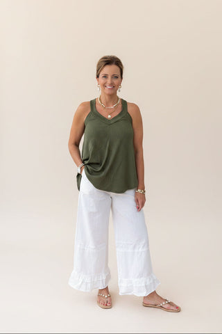 Woman wearing an olive green sleeveless top and white pants against a beige background
