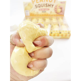A hand squeezes a yellow, peanut-shaped squishy toy, with a blurred box of Peanut Squishy candy in the background.