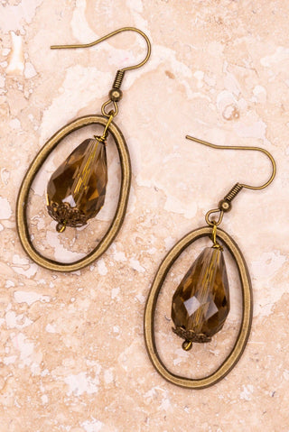 A pair of dangling earrings with oval bronze-toned metal frames and faceted brown teardrop gemstones, displayed on a light beige stone-textured background.