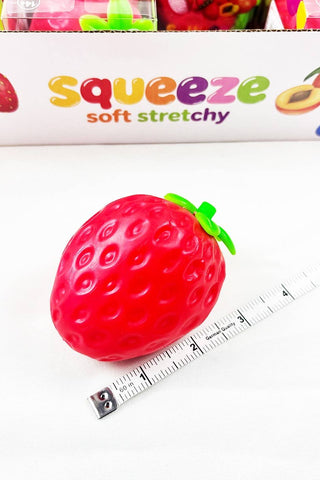 A red strawberry-shaped squishy toy with a green stem, shown next to a measuring tape and in front of its product packaging box labeled 'squeeze soft stretchy'.