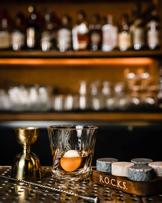Bar setup with a glass of whiskey, cocktail shaker, and 'R.O.C.K.S.' container against a blurred bar background.