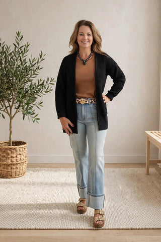 A woman stands in a bright, neutral room wearing a black cardigan over a brown top, light blue jeans, and brown sandals. She is posed in front of a beige wall with a potted plant on the left and a wooden bench on the right.