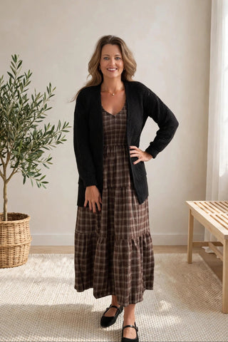 A woman stands indoors modeling a long brown and beige plaid dress with a black cardigan, black flats, in a bright room with a potted plant and a wooden bench.