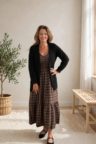 A woman stands indoors modeling a long brown and beige plaid dress with a black cardigan, black flats, in a bright room with a potted plant and a wooden bench.
