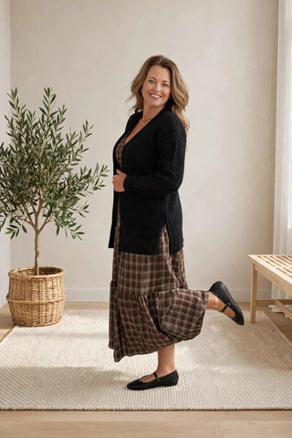 A woman modeling a black cardigan over a brown and beige plaid maxi skirt, standing in a bright, minimalist room with a potted olive tree and a wooden bench.