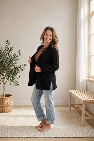 A woman stands barefoot in a bright room wearing a black open-front cardigan over a brown top and light blue jeans.