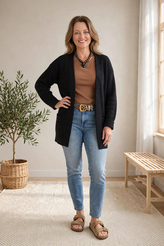 A woman modeling a black ribbed cardigan over a brown top, blue jeans, and tan sandals in a bright, neutral indoor room with a potted plant and wooden bench.