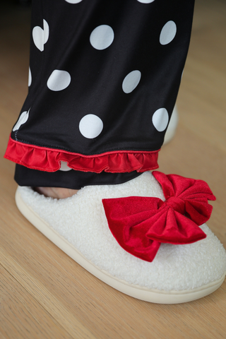 White slippers with red bows worn with black pajamas with white polka dots with red ruffle on a wooden floor.