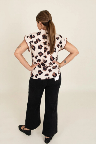 A woman is shown from behind wearing a short-sleeve top with a black and brown leopard print on a light pinkish-beige background, paired with black wide-leg pants and black shoes, against a plain light beige studio background.
